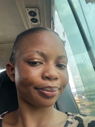 A portrait-style selfie features a young adult with dark skin and very short, dark hair, possibly a buzz cut. The subject looks directly at the camera with a calm, smiling, or smirking expression, showcasing full, glossy lips and a small gold stud earring in their left ear. The image is notably rotated 90 degrees counter-clockwise, with the subject's head tilted to the left.
The setting is inside a vehicle, likely a car or bus, during daytime in Satellite Town, Nigeria. Above and behind the subject's head, a large, clear or lightly tinted window reveals a blurred outdoor scene with green foliage from trees and indistinct buildings or structures, suggesting an urban or suburban environment. The vehicle's interior is visible, with dark grey or black surfaces around the subject's shoulder and neck. To the left, a distinct light-colored panel holds a rectangular unit containing two circular speakers. The subject is wearing a patterned top, possibly an animal print, in black and light brown/cream, visible on the right side of the frame. The natural daylight indicates daytime. No text is visible in the image.