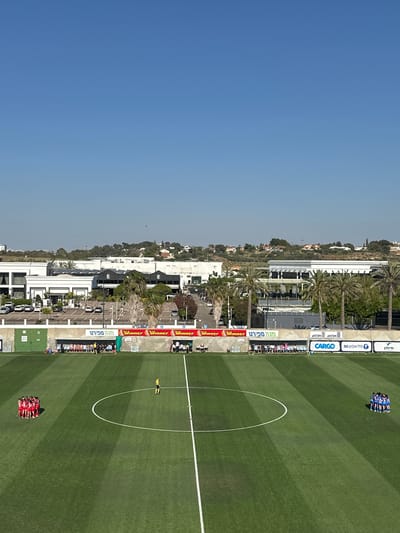 This aerial shot depicts a soccer field in Ness Ziona, Israel, juxtaposed against the city skyline. On the right, a lush green field features white lines marking its boundaries, including a prominent center circle. Two groups of soccer players, one in blue and the other in red, are gathered in huddles near opposite ends of the field. Along the sideline, advertising banners display brands such as "Mega Sport", "Winner", "Cargo", "Mei Eden", and "Pelephone 5G" in Hebrew.

Beyond the field, the urban landscape of Ness Ziona unfolds with residential buildings, palm trees, and roadways. Cars are parked along the street. The left portion of the image is dominated by a clear, light blue sky.