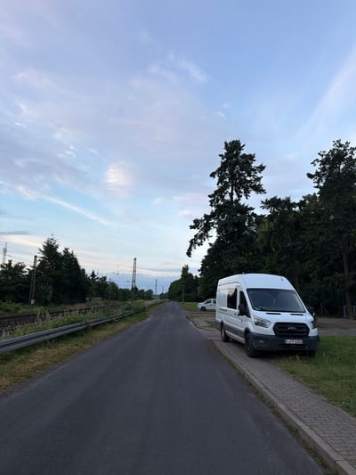 A white van, identified by its license plate "F-FP 2353," is parked on the right side of a paved road in Magdeburg, Germany. The van is facing forward, towards the distance. To the left of the road, train tracks run parallel, bordered by a metal guardrail and overgrown vegetation. In the background, tall trees line the right side of the road, creating a dense green backdrop. The sky above is a pale blue with scattered wispy clouds, suggesting either early morning or late evening. The overall environment appears to be on the outskirts of the city, possibly near a transportation hub or industrial area.