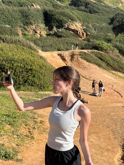The image portrays an outdoor scene on a sunny day in Palos Verdes Estates, United States, featuring a network of reddish-brown dirt paths winding through hills densely covered with green shrubbery and some exposed rock formations. In the foreground, a young Caucasian female with brown hair styled in a side braid, wearing a light grey ribbed tank top over a dark sports bra and black athletic shorts, extends her right arm horizontally. She holds a black smartphone in her right hand, angled slightly upward, indicating a recording or photographic action. Her gaze is directed to the right of the frame. In the midground, further up the dirt path, two females in casual or athletic attire are visible, one in a black top and light shorts, the other in a white top and light shorts, appearing to stand or walk. A light-colored object, possibly a bag, rests on the ground near them. Higher on a distinct winding dirt path in the background, two smaller figures, one wearing a pink hat, are observed walking away from the viewer.