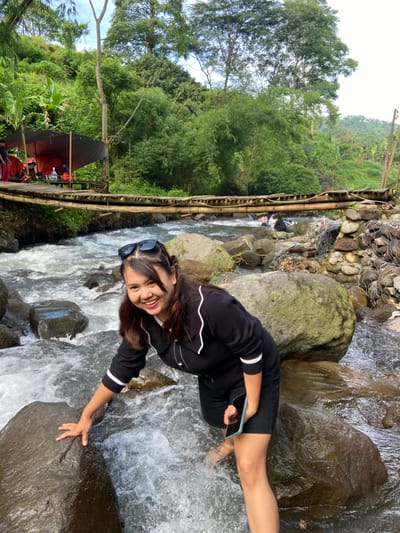 A young woman kneels by a rocky stream, her hand resting on a large stone. She wears a black, long-sleeved shirt with white stripes on the sleeves, and black shorts. Sunglasses rest on her head, and she smiles at the camera. A phone is in her hand. Lush green trees surround the area, with a wooden structure and red umbrellas visible in the background. The water rushes over the rocks, creating a dynamic and natural setting.