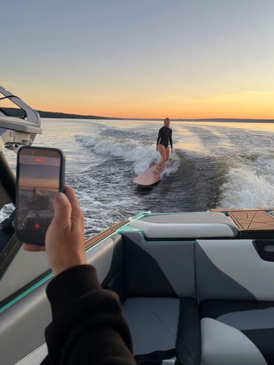 An outdoor, eye-level, medium shot captures a person holding a smartphone in the foreground, filming a woman wake surfing on a lake during sunset. The phone is held in a person's right hand and arm, with the phone screen displaying a recording of the scene. The woman in the background, wearing a black wetsuit, is actively surfing a wave behind a boat, which is partially visible in the lower right corner of the frame. The lake is calm, with a gentle sunset casting a warm orange glow across the sky. Trees line the distant horizon, suggesting a natural landscape surrounding the lake. The water is dark, with white foam from the wake of the boat and the surfing activity. The overall atmosphere is serene and active, highlighting the combination of outdoor recreation and personal technology. The location is Gromovo, Russia.