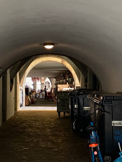 An interior view shows an arched passageway in Lucerne, Switzerland. The shot is framed by the rounded archway, which is lit by a small wall sconce to the left. In the distance, a series of arches lead to a bright, sunlit window at the far end. People are seated in the space beyond, possibly reading. Stacked black cases and a blue bicycle lean against the right side of the archway. The floor is paved with light-colored stones.
