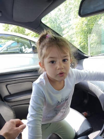 A young girl is in the front seat of a car. The girl has brown hair, fair skin, and is wearing a white long-sleeved shirt. The car's interior is grey and black. The image is taken from a slightly high angle looking down at the girl. Outside the car, green trees and foliage are visible. There is no explicit indication that the picture was taken in Stafford, United Kingdom.