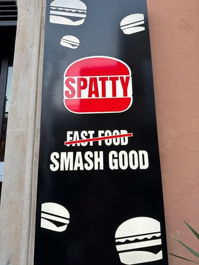 A sign for "SPATTY" reads "FAST FOOD SMASH GOOD" with "FAST FOOD" crossed out. The sign is black with white text and red logo. The sign has burger icons along the edges. The location is Acireale, Italy.
