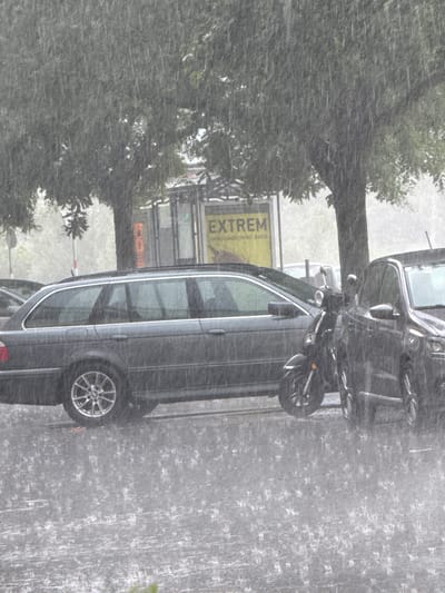 The image depicts a street scene in Innsbruck, Austria, during a heavy downpour. Rain is visible as streaks across the frame, with many splashes on the asphalt ground, indicating active precipitation.

In the foreground, a dark-colored station wagon is parked, facing left. Its wheels are visible, and the car is wet from the rain. To the right of the station wagon, a black scooter is parked, also appearing wet. Beside the scooter, the front of a dark-colored car is partially visible, facing right.

Behind the vehicles, a tree with dense green foliage is prominent, its branches partially obscured by the rain. To the left of the tree, a light pole with a circular sign is present. Behind the tree and station wagon, a yellow billboard is visible with the word "EXTREM" in large letters. Further back, a bus stop shelter with a glass facade can be seen. The overall environment appears to be an urban street or parking area, with trees lining the sides. The weather conditions are clearly severe, with the rain creating a blurred and wet atmosphere. There are no people visible in the image.