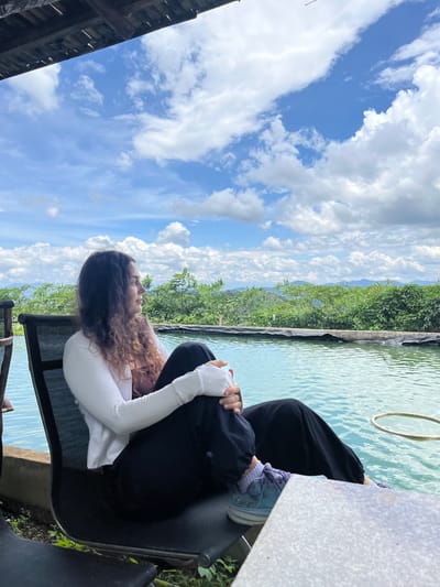 A woman with long, curly brown hair sits in a black reclining chair next to a pool of blue water. She is wearing a white long-sleeved shirt, a brown camisole, and black pants. Her legs are crossed, and she is looking off to the right. The pool is surrounded by lush green vegetation, and in the background, there are mountains under a bright blue sky with white clouds. The scene appears to be an outdoor relaxation area. The location is in Son Dien city, Vietnam.
