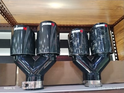 This media file displays a close-up, eye-level shot of four exhaust tips, two on the left and two on the right, mounted on a shelf. The exhaust tips are constructed from carbon fiber and black metal. The left pair features a black carbon fiber pattern with a red "AKRAPOVIĆ" logo on each tip. The right pair has a different carbon fiber pattern, also with the red "AKRAPOVIĆ" logo. Each set of exhaust tips has a Y-shaped black metal connector with a silver clamp at the bottom of each. The background consists of two light brown cardboard boxes, and a wooden shelf with metal framing. The lighting suggests an indoor setting, possibly a store or workshop. Visible text includes "REDMI 15" in the bottom left corner and a date and time stamp "22.06.2026 17:03" in the bottom right corner. There are no people or animals present, and no weather or time of day cues are discernible other than the timestamp. The location is identified as Yovon, Tajikistan.