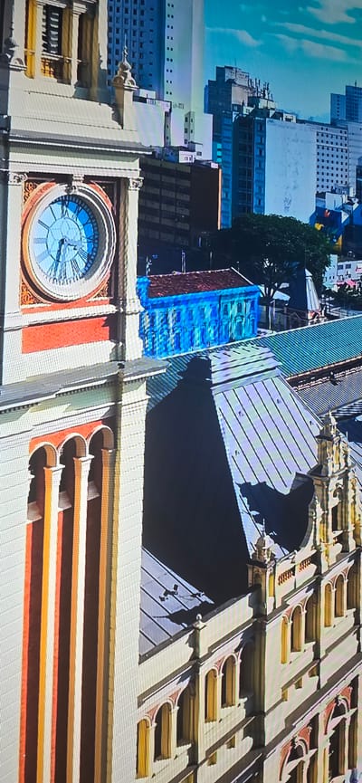 The image shows a close-up, aerial view of an ornate building with a clock tower, likely in Lippstadt, Germany. The clock face is visible, showing the time as approximately 10:55. The building features arched windows and decorative columns. In the background, modern city buildings can be seen, along with a tree and a bright blue structure. The lighting suggests it is daytime. There are no people visible in the frame.