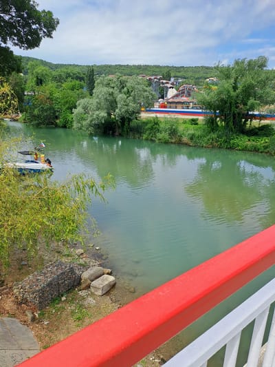 The image shows a view of a river in Dzhubga, Russia. In the foreground, the red and white railing of a bridge is visible. The river itself is calm and a greenish-blue color. On the left bank of the river, a small white boat with a blue stripe is docked, and a person wearing an orange life vest is standing on it. There is also a Russian flag on the boat. The bank is lined with rocks and vegetation, including a weeping willow tree.
In the background, across the river, there is a water park with several colorful slides and structures. The area is surrounded by lush green trees and a hillside. The sky is partly cloudy, with patches of blue sky visible. The overall scene depicts a recreational area by the river.