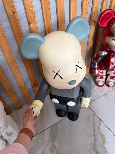 A person's arm and hand is visible in the lower left of the frame, wearing a brown watch and a pink shirt sleeve. The hand is reaching out to touch the foot of a large, gray and cream-colored toy figure with blue ears and black X's for eyes. Behind the toy figure is a red and silver toy figure. The figures are standing on a marble-patterned floor, in front of a wall made of vertical wooden planks and gray paneling. The scene is indoors, likely in a residential setting or shop in the city of Semiluki, Russia.