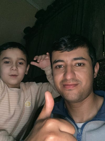 A low-light indoor photograph taken in Galaosiyo, Uzbekistan, depicts two individuals. In the foreground, an adult male, appearing to be of Central Asian descent, with short dark hair and stubble, wears a grey zip-up jacket with a blue collar. He looks towards the camera with a slight smile and extends his right hand into the foreground, forming a thumbs-up gesture. To his left, partially behind him, a male child, approximately 6-10 years old, also of Central Asian descent, wears a light-colored, possibly cream or beige, horizontally striped long-sleeved shirt with a small circular yellow citrus fruit logo on the chest. The child looks at the camera with a neutral expression, his left hand raised near his head with fingers slightly curled. The background is dark and indistinct, revealing shadowed shapes suggestive of indoor furnishings.