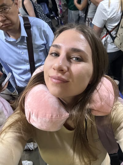 A female individual is depicted in a close-up, selfie-style photograph within a crowded indoor public space, identified as potentially Phuket International Airport in ตำบล ไม้ขาว, Thailand. The subject, appearing to be in her 20s or 30s, has long brown hair, light skin, and green eyes, and displays a subtle smile towards the camera. She wears a light-colored, long-sleeved top and a pink velvet U-shaped travel neck pillow. A dark brown backpack strap is visible over her right shoulder. Behind and to the left of the main subject, a male with glasses, wearing a light blue collared shirt, is partially visible, carrying a dark brown messenger bag with "BOSTA" printed on its strap. His expression is neutral. Further background elements show additional indistinct individuals and partial views of luggage, consistent with a busy travel terminal environment. The lighting is artificial and bright.