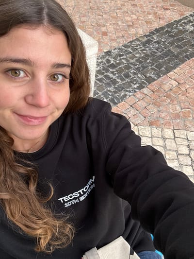 A young adult female with long brown hair and green eyes is depicted in a close-up, high-angle selfie. She wears a black sweatshirt with "TECSTORM 10TH EDITION" printed in white on the chest and a delicate gold necklace. Her lips are slightly parted in a smile. Behind her, a segment of a light-colored stone structure is visible. The background consists of a patterned cobblestone pavement, featuring diagonal stripes of dark grey stones alternating with reddish-brown stones. The scene takes place outdoors in Porto Salvo, Portugal.