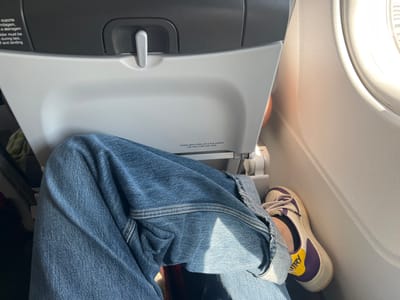 The image captures a close-up view of a person's legs and feet, clad in blue jeans and colorful sneakers, inside an airplane cabin. The perspective is from the point of view of the person, looking down at their lap and extending towards their feet. The airplane seat in front of them is visible, with some text printed on its surface.

The seatback in front has visible text. One section reads "suporte rolagem, e aterragem" and "alder must be during taxi, and landing". Another section has the text "RECARO" on a small lever. Further down on the seatback, more text reads "Colete salva-vidas sob a sua cadeira" and "Life vest under your seat". The lighting suggests it is daytime, with sunlight streaming in from a window on the right side of the frame, creating bright highlights and soft shadows on the seat and the person's clothing. The airplane interior appears to be modern and functional, typical of a commercial flight.

The person is wearing a white sneaker with purple and yellow accents. The jeans are a classic blue denim, with visible stitching and folds, indicating the relaxed posture of the person. The overall scene conveys a sense of travel and the ordinary experience of being on an airplane. There are no explicit emotions conveyed, but the focus on personal space and comfort, indicated by the relaxed leg posture, suggests a typical passenger on a journey. The location context of Paray-Vieille-Poste, France, implies this is likely a flight departing from or arriving at Paris-Orly Airport.