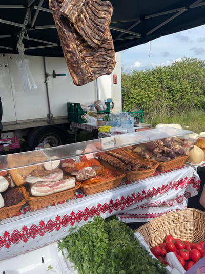 The image shows a vendor stall, likely at a market. In the foreground, a large piece of dried, cured meat hangs from the stall's frame. Behind it is a white truck with what appear to be various food items displayed in a clear case. Baskets of cured meats and cheeses are visible within the case. A white cloth with a red embroidered pattern is next to the clear case. Baskets of tomatoes and bunches of parsley are also visible. The background includes green trees and a cloudy sky. The location is unknown.