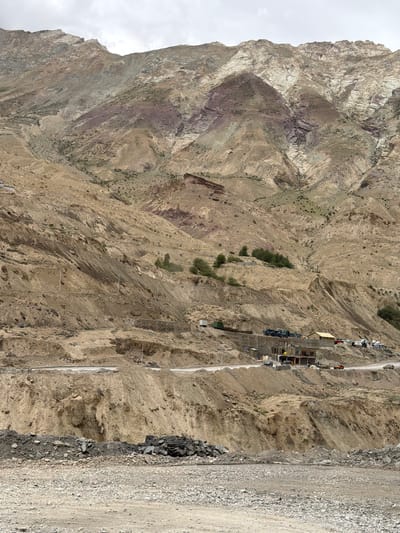 A wide, arid mountain landscape dominates the scene. The mountains are characterized by their rugged, eroded slopes, displaying layers of tan, brown, and reddish-purple rock. Sparse green vegetation clings to some of the lower slopes. A road winds through the mid-ground, carved into the mountainside, with evidence of construction activity. Several buildings, some appearing to be under construction and others more established, are visible along the road. Various vehicles, including trucks, cars, and possibly construction equipment, are parked or in motion near the buildings. The sky is overcast, suggesting a cloudy day. The foreground is covered in loose gravel and rocks.