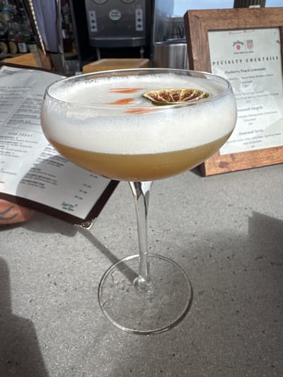 An outdoor table at a restaurant in Marina del Rey, United States, features a coupe glass filled with a foamy, amber-colored cocktail. The drink is garnished with a dried citrus slice and a few small red edible flowers. To the left of the glass, a menu lies open, and to the right, a framed menu with the words "SPECIALTY COCKTAILS" is visible. In the background, a bar area with refrigerators and bottles can be seen. The table surface is a gray, speckled material.