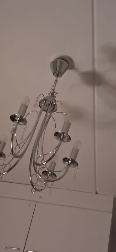 A silver chandelier with crystal accents hangs from a white ceiling. The chandelier has multiple arms, each ending in a light socket designed to resemble a candle flame. Several bulbs are visible, and the chandelier is adorned with dangling crystal elements. Below the chandelier, the top of white cabinets with silver handles can be seen. The scene is illuminated, casting shadows from the chandelier onto the ceiling and an adjacent wall, suggesting indoor lighting. The image composition is a close-up, angled shot of the chandelier and its immediate surroundings. There are no people or animals visible in the frame. The image appears to have been taken in Lahti, Finland, within a domestic or commercial interior space.