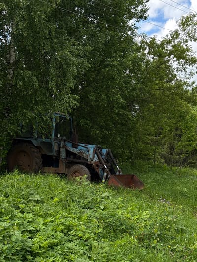 The image depicts a derelict, light blue tractor partially obscured by dense vegetation. The vehicle, showing signs of rust and disuse, is positioned in an overgrown area with tall grass and trees. Power lines are visible in the upper left corner of the image. The location is purported to be Medvedka, Russia. No people are present.