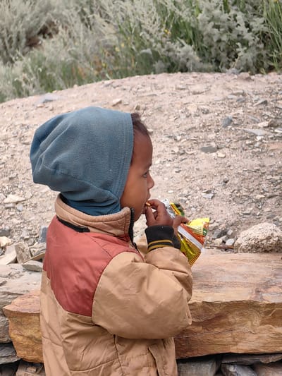 A child, seen in profile and from the chest up, sits on a large rock in a dry, rocky environment with sparse vegetation in the background. The child wears a blue fleece hood pulled over their head and a tan and reddish-brown puffy jacket with a black and yellow striped cuff. They are holding a yellow and red bag of chips with their right hand and eating from it with their left. The scene is likely in or around Gondhla, India, given the arid landscape.