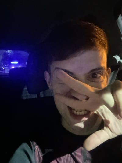 A man is shown in a distorted, low-lit selfie, likely taken at night. His face is illuminated by a warm light source, contrasting with the dark background. He partially covers his face with one hand, drawing attention to his eye and teeth. The background includes hints of blue light, suggesting an urban environment. The overall image has a candid, slightly unsettling feel.