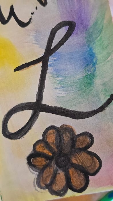 The image presents a close-up, cropped view of a piece of artwork, appearing to be watercolor on paper with black ink or marker detailing. The background consists of blended watercolor washes arranged in horizontal bands: yellow in the lower left, transitioning to light blue, then green, and finally purple and pink towards the upper right. Prominently featured in the foreground is a large, black, stylized cursive uppercase letter "L," extending across the central and right portions of the frame. Above and to the left of the "L," a partial black cursive character, resembling the upper curve of a "w," is visible. Below the "L" and slightly to the left, a stylized flower is depicted with eight rounded petals rendered in brown with a distinct black outline, and a dark, solid black center. The paper surface shows subtle texture. No people, specific actions, or interactions are visible. The scene is located in Lompoc, United States.