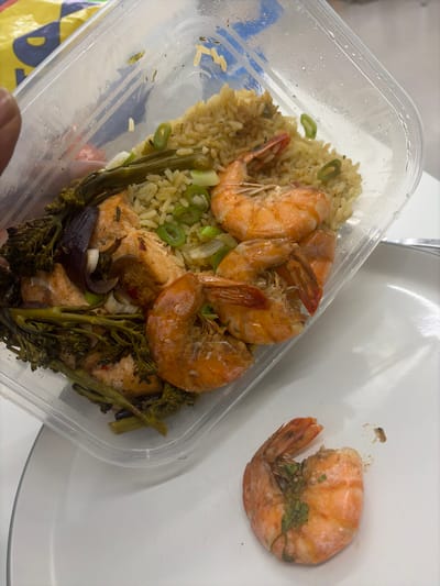 A clear, rectangular plastic food container is held by a human hand, primarily visible as a thumb and index finger, in the upper-left of the frame. The container holds a prepared meal consisting of light brown rice, several cooked, orange-colored prawns (some partially peeled with tails attached), light pink pieces of cooked salmon, dark green broccolini florets, and slices of purple onion. Green chopped scallions are distributed among the food items. Condensation or moisture is present on the interior surface of the container.
Adjacent to the container, resting on a white, flat surface, is a single, cooked, partially peeled prawn with its tail segment still attached. A small, dark, granular food particle is located near this detached prawn. The white surface appears to be a table or countertop. A faint, curved line suggests the presence of a white plate partially obscured by the container. In the blurred background, a yellow and blue object is partially visible. The scene is illuminated by ambient light. This activity is occurring in New Forest, United Kingdom.