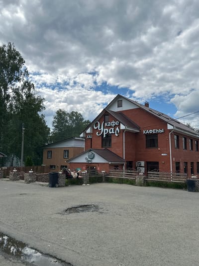 The image shows a brick building with a sign that reads "КАФЕ УРАЛ". In front of the building is a dirt road with several potholes. A person is lying on a bench in front of the building, seemingly asleep. The sky is cloudy. The location is the city of Inzer, Russia.