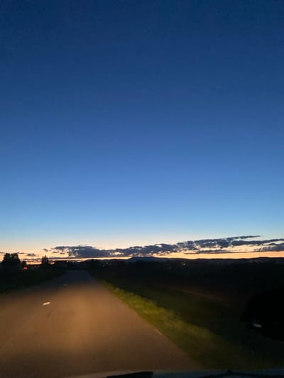 The image captures a rural road scene near Hurtigheim, France, during twilight or early night. The sky, occupying approximately two-thirds of the frame, transitions from a deep indigo blue overhead to a lighter blue-orange gradient at the horizon. A horizontal band of dark, elongated clouds is present directly above the horizon line. Below the horizon, distant dark hills or mountains are discernible, beneath which faint artificial lights from a settlement are visible.

A two-lane asphalt road extends from the foreground into the midground, illuminated by a vehicle's headlights, which cast a yellow-orange light forward onto the road surface and the immediate grassy verges. White dashed lane markings are visible on the asphalt. The right side of the road appears bordered by dark fields. The perspective suggests the image was captured from inside a vehicle, looking forward. No individuals or overt actions are present in the scene.