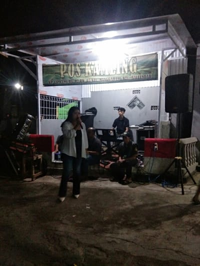 The image captures a nighttime outdoor performance or community gathering under a temporary, open-sided structure, likely a canopy, supported by metal beams. The scene is brightly lit by overhead artificial lights, contrasting with the dark surroundings.

In the center foreground, a woman stands holding a microphone to her mouth, appearing to sing or speak. She is wearing a light-colored (possibly white) long-sleeved shirt over a dark top, dark trousers, and light-colored pointed shoes.

Behind her, several men are positioned as if part of a band or accompanying musicians. To her right (viewer's left), a man is seated, partially obscured. Further back and to the right, another man is seated at a keyboard or synthesizer, seemingly playing the instrument. Another man is visible seated on the right side of the stage area. On the far left, a stand that appears to be for a drum set or other percussion instrument is visible.

The performance area is set up against a light-colored wall or building, which features some decorative or ventilation blocks in its upper section.

Above the stage, a prominent green banner hangs, displaying white text. The main text reads "POS KAMLING". Below this, in smaller white letters, it states: "KP. CIPICUNG RT. 02 RW. 03 KEL. MANGGAHANG KEC. BALEENDAH".

Equipment for the performance is visible around the stage. On the far right, a large black speaker is mounted on a stand. Closer to the ground on the right, a smaller red and white speaker or amplifier is placed, surrounded by various cables.

The ground surface in the foreground appears to be dark, possibly concrete or pavement, showing some texture. The overall atmosphere suggests a casual, local event or performance taking place during the evening.
