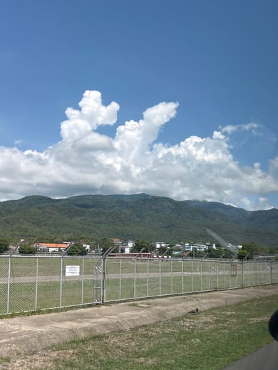 A photograph depicts a landscape in Suthep, Thailand. The sky, predominantly blue, is punctuated by fluffy white clouds. A large, green mountain dominates the midground, its slopes covered in dense trees. In the foreground, a chain-link fence runs parallel to a concrete path, with a grassy area on the other side. Beyond the fence, there are several buildings and vegetation. A bird stands by the concrete path.