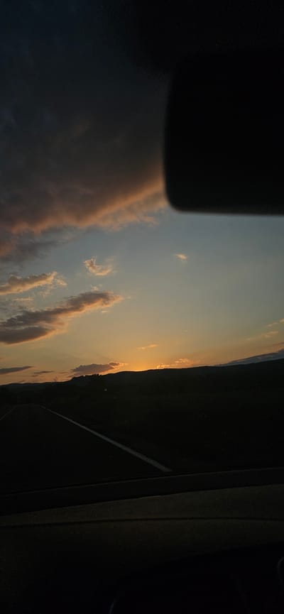 A horizontal, wide-angle photograph, taken from the interior of a vehicle, captures a sunset over a rural landscape in the vicinity of Dolna Banya, Bulgaria. The upper portion of the sky transitions from dark, heavy clouds with grey and subtle pink hues in the upper-left, to a clear, pale blue in the upper-mid, then to an intense orange and yellow glow at the horizon, indicative of a sunset. Scattered, elongated cirrus clouds, reflecting warm orange and pink light, are present across the mid-sky. A faint, slender crescent moon is visible in the pale blue section, slightly right of center.
The sun itself is positioned behind a distant, silhouetted mountain range, creating a bright orange luminosity just above the dark peaks. Below the mountain range, the mid-ground landscape is rendered in deep silhouette, suggesting dense terrain or fields. In the lower-left foreground, a dark asphalt road is visible, marked by a white dashed line receding towards the center. The immediate foreground shows the dark dashboard of the vehicle, obscuring the bottom of the frame. A large, dark, irregular shape, consistent with an automotive component (e.g., rearview mirror or sun visor), occupies the upper-right quarter of the image, partially obstructing the sky. No individuals or direct interactions are visible.