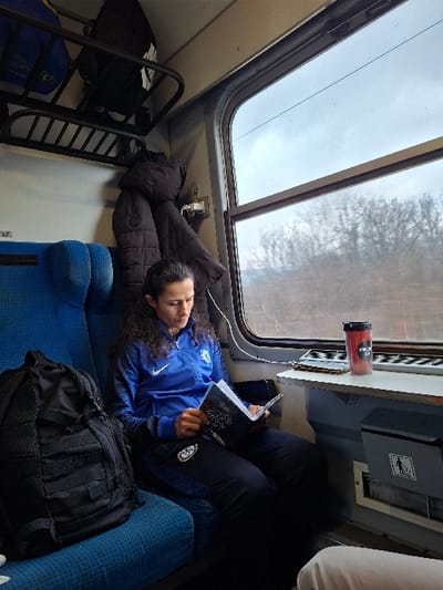 An interior view of a train compartment, likely in motion near Lyutibrod, Bulgaria, shows a female passenger seated on a blue upholstered seat on the left side of the frame. She has dark, curly hair pulled back and is wearing a blue athletic jacket with a white Nike logo and a Chelsea FC crest, along with dark athletic pants featuring a similar crest. She is focused on reading an open book with a dark cover. To her immediate left, on the same seat, a black tactical-style backpack rests. To her right, a small grey table attached to the window frame holds a red and black travel mug with a black lid and white text. Beyond the window, a blurred landscape of bare trees and an overcast sky is visible, indicating movement. Above the passenger, an overhead luggage rack contains a blue bag and a black bag. A dark brown padded jacket hangs from a hook below the luggage rack. A grey waste receptacle with a universal trash icon is located beneath the table. The lower right corner of the frame reveals a portion of a light-colored pant leg, suggesting the presence of another passenger out of view.