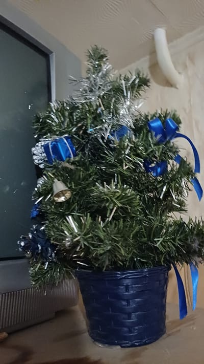 The image shows a small, artificial Christmas tree decorated with blue bows, silver tinsel, small blue wrapped gifts, and a single gold bell. The tree is in a dark blue, woven pot. The tree is situated on a surface, possibly a shelf or table, next to a dark-colored television or monitor. The background consists of a textured wall and ceiling, with a white pipe or conduit visible on the wall. The lighting suggests an indoor setting, likely a home or office environment. There are no people visible in the image. The specific location in Murmansk, Russia cannot be determined from the visual information provided.