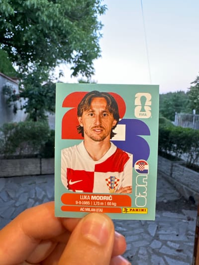 A hand holds a collectible trading card featuring soccer player Luka Modric. The card has a light blue background with a stylized image of Modric in his Croatian national team jersey. Text on the card includes his name, birthdate, height, weight, and club (AC Milan). The card also features the FIFA logo and the Panini logo. In the background, there is greenery and a hint of a building, suggesting an outdoor setting. The location is identified as the city of Prisovo, Bulgaria.