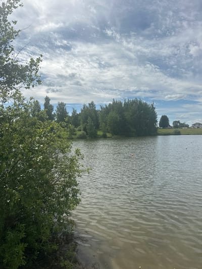 The image shows a body of water with rippling surface, reflecting the sky. The sky is filled with scattered clouds, some wispy and others more dense. On the left side of the image, a line of trees and lush green bushes runs along the bank of the water. A few smaller trees are visible further back, and in the distance, a building with a white facade and dark roof can be seen. Two small orange buoys are floating on the water. The scene appears to be a natural landscape, possibly a lake or a wide river, under a partly cloudy sky. The overall impression is one of a serene and natural environment, likely captured during daylight hours, given the brightness and cloud formations. There is no visible text. The location context suggests this is in the Пярэжырскі сельскі Савет region of Belarus.