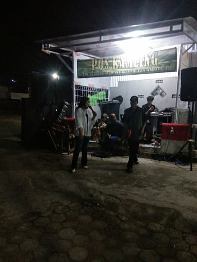This nighttime image captures an outdoor community gathering or performance in Baleendah, Indonesia. A woman, prominently featured in the foreground, wears a white jacket and dark pants, holding a red microphone as if singing or addressing the crowd. To her right, a man in a dark shirt and cap is seen walking, while another man operates sound equipment on a slightly raised platform. Several other individuals are visible in the background, some seated.

The event takes place under a simple, open-sided shelter illuminated by overhead lights. A large banner stretches across the front of the shelter, displaying the prominent text "POS KAMLING" (Community Security Post). Below this, smaller text specifies the location: "KP. CIPIJUNG RT. 02 RW. 03 KEL. MANGGAHANG KEC. BALEENDAH," indicating a specific hamlet and administrative details within the Manggahang village of Baleendah district.

Visible objects include a large black speaker on a stand to the right, a sound mixer or DJ setup on a table, and a red and white equipment box. To the left, behind the woman, what appears to be a drum or percussion instrument is partially visible. The ground is paved, suggesting a public or communal space. The scene conveys a casual and engaged atmosphere, indicative of local entertainment or a community function.