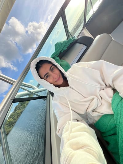 The image displays a female individual taking a selfie inside a boat on a body of water, specifically identified as the Minho River near Vila Nova de Cerveira, Portugal. The subject, with dark hair and light eyes, is positioned centrally, looking directly at the camera with a subtle smile. She is wearing a light-colored, possibly off-white, hooded zipper sweatshirt with the hood raised. Her left arm is extended towards the camera. To her immediate right, a light-colored upholstered boat seat is visible, along with a bright green towel or fabric draped over a surface. The boat's structure, including a glass windshield and metallic framing, is prominent around her. Beyond the boat's interior, a partially cloudy blue sky is visible above. The water outside the boat is calm with visible ripples, reflecting the sunlight. Distant, tree-covered hills line the horizon across the water.