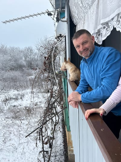 A forensic examination of the image reveals a winter scene captured from a balcony in Synelnykove, Ukraine. The foreground features a male individual, observed from the chest up, positioned towards the right of the frame. He has short, dark hair and a beard, and wears a blue fleece half-zip shirt. He is smiling and looking directly at the camera. His forearms rest on a dark brown wooden balcony railing. To his right, a female hand with red-painted nails, adorned with a light pink ribbed sweater cuff, also rests on the railing. To the man's left, a light brown dog with shaggy fur, possibly a terrier breed, stands on the railing, looking intently towards the snowy outdoor landscape.
The immediate balcony environment includes white vertical siding along the inner wall. Above, a white lace curtain is partially visible at the top right. Directly above the balcony, an outdoor Yagi antenna and a circular satellite dish, both appearing to be coated with ice or snow, are mounted. Bare, dark vines with remnants of dry leaves or berries cling to the structure supporting the balcony, extending downwards into the outdoor scene.
The exterior environment is characterized by a thick blanket of white snow covering the ground and the numerous bare branches of deciduous trees and bushes. The sky is overcast and bright. The overall impression is a cold, snowy day. In the middle distance, hints of a reddish-brown structure are faintly visible amidst the snow-laden trees.