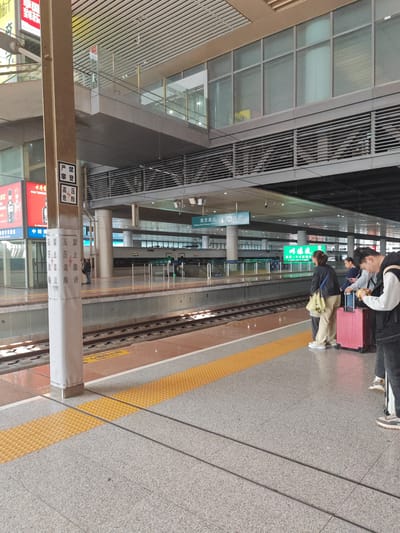 This image depicts an active platform at Nanjing South Railway Station, located in Yuhuatai District, China. The scene showcases a modern indoor train station with a high, paneled ceiling, elevated structures featuring glass windows and ventilation grilles, and tracks running alongside the platform.
In the foreground, a sturdy square pillar displays multiple safety warnings in Chinese, including "严禁 攀登" (Strictly forbidden to climb), "高压 危险" (High voltage, danger), and a repeated vertical banner stating "高压危险 严禁靠近" (High voltage danger, strictly forbidden to approach). The platform floor is a speckled grey, with a bright yellow tactile paving strip marking the edge adjacent to the tracks.
Several people are visible on the platform, engaged in typical waiting or transit activities. In the mid-ground right, three individuals are gathered; two women, seen from behind, with one carrying a yellow bag and another standing beside a pink rolling suitcase. A man next to them is looking down, likely at a phone. Other passengers and possibly staff are scattered further down the platform. A train is partially visible on the tracks in the background.
Overhead, a blue sign clearly indicates "南京南站" (Nanjing South Station) and its English equivalent, "Nanjing South Railway Station," along with an "出站" (Exit) arrow. Another sign denotes "中转" (Transfer). On the left, an advertisement for "古井贡" (Gujinggong), a brand of Chinese liquor, is displayed. The station is well-lit by artificial lights, suggesting daytime or consistent interior illumination. The atmosphere appears calm and orderly, characteristic of a busy transport hub.