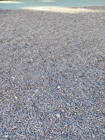 This media file depicts a close-up, eye-level view of a ground covered in small, multi-colored gravel. The gravel is primarily composed of shades of gray, brown, and some reddish-pink hues, with individual stones appearing rough and angular. In the upper portion of the image, a paved area with a distinct pattern of rectangular blocks is visible, illuminated by sunlight. This paved area transitions into the gravel ground.
The scene suggests an outdoor setting, likely a pathway, courtyard, or garden area. The lighting indicates it is daytime, with bright sunlight casting a sheen on the paved surface and creating dappled shadows or highlights across the gravel. The overall impression is one of a dry, possibly sunny, day. There is no discernible activity or event taking place; the focus is solely on the texture and composition of the ground. The location context of Kaluga, Russia, does not provide specific visual cues within the image itself, as the gravel and paving are common landscaping materials. No text is visible in the media file.