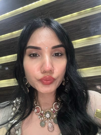 A close-up self-portrait depicts a woman with long, dark, wavy hair, looking directly at the camera with her lips pursed. A small, clear jewel is affixed to her forehead between her eyebrows. She wears elaborate jewelry, including a prominent gold-toned necklace with circular and teardrop elements set with green, pink, and clear stones, and matching dangling earrings. Her attire, partially visible on her left shoulder, is a light-colored fabric adorned with sequins and pearls. The background features horizontal striped paneling, alternating dark textured bands with illuminated lighter bands. The specific location in New Town, India, cannot be determined from the visual information provided.