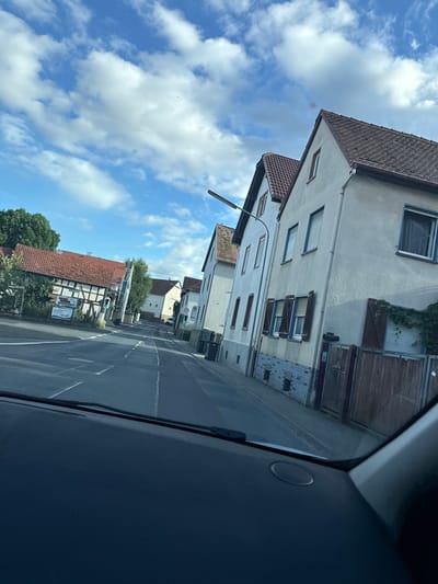 The image depicts a view from a vehicle driving down a street in Weidenhausen, Germany. The street is paved with asphalt and marked with white lines indicating lanes. On the right side of the street are several residential buildings with light-colored facades, red tile roofs, and windows. Some of the buildings have wooden shutters and fences. On the left side of the street, there is a building with a half-timbered facade, a red tile roof, and a sign with German text and an image. Trees and foliage are visible behind this building. The sky is blue with scattered white clouds. Traffic lights are visible, with one showing a green light. Several trash bins are present on the side of the road. The dashboard of the vehicle is partially visible in the foreground.