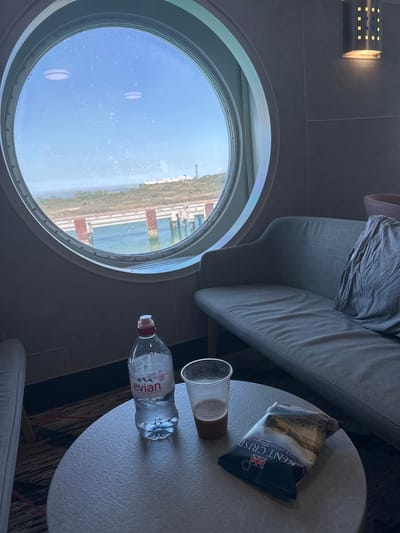The image depicts an interior scene, likely aboard a ferry or ship, as evidenced by a circular porthole window offering a view of what appears to be the coastline of Loon-Plage, France. The view through the window shows a blue sky with some cloud cover, and land with vegetation along the shoreline. Man-made structures are also visible, including rectangular buildings and other architectural features.
Inside the space, there is a grey sofa with a crumpled grey fabric on it. Next to the sofa, there is a small round table. On the table, there is a bottle of "Evian" water, an empty drinking glass, and a package of "Kent Crisps" sea salt and vinegar potato chips. A light fixture is visible on the wall.
