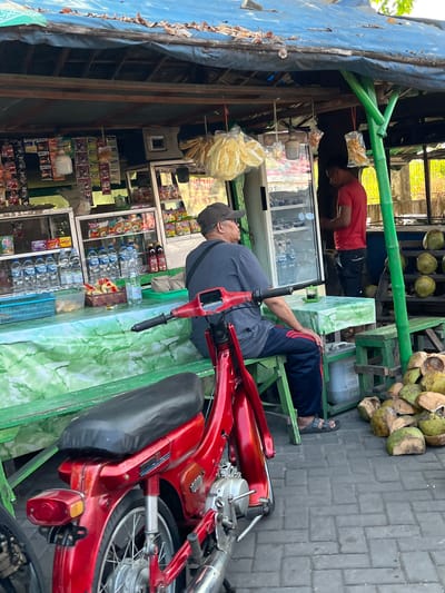 The image depicts a street-side refreshment stall in Juwet Kenongo, Indonesia. A man wearing a dark blue t-shirt and dark pants with red stripes sits on a green bench in front of the stall. He is facing away from the camera, looking towards a small table with a cup of dark liquid. Beside him, a red motorcycle is parked. Behind the man is a stall with various items for sale displayed on shelves, including bottled water, snacks, and other packaged goods. A young man in a red t-shirt stands inside the stall, near a refrigerator unit. A stack of halved coconuts is visible on the right side of the image, near a wooden stand. The stall is covered by a blue tarp. The ground is paved with grey interlocking bricks.