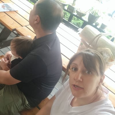 The image captures a family selfie taken outdoors at a picnic table, possibly in Mureungdowon-myeon, South Korea. In the foreground, a woman is looking at the camera with an open mouth. To her left, a baby is being held by a man, whose back is mostly to the camera. The man is wearing a dark shirt and green pants. Behind them, a picnic table is visible, with a magazine or book on it. In the background, there are plants in pots on a railing, suggesting an outdoor seating area or balcony. The lighting is bright, indicating it is daytime. The overall scene depicts a casual family moment.