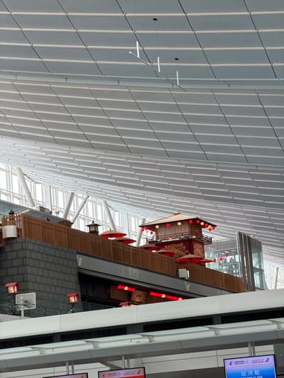 The image shows the interior of a modern building, likely an airport terminal in Ota, Japan, with a distinct Japanese-inspired architectural element. The ceiling is a complex, multi-layered structure of white, diamond-shaped panels, interspersed with light fixtures. Below the ceiling, a multi-story, wooden structure with traditional Japanese architectural features is visible. This structure is adorned with red lanterns and decorative elements, including red umbrellas. The facade of the lower levels features a dark, textured material resembling brick, topped by wooden railings and panels. Several windows and a modern glass elevator shaft are also present in the background, suggesting a public space. The overall impression is a blend of contemporary design and traditional Japanese aesthetics.