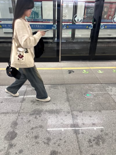 A person with long black hair stands on a gray tiled platform, looking at their phone. They wear blue jeans, white sneakers, and a beige sweater with cartoon patches. A black backpack is slung over their shoulder, and a black cap hangs off their bag. A yellow line with green arrow signs runs along the platform edge next to subway doors. Chinese characters are visible on the train doors and platform information boards.