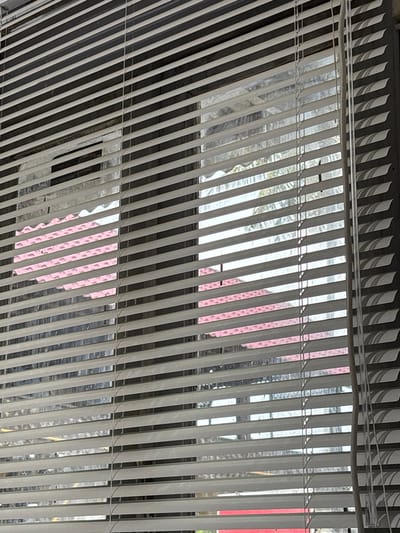 The image shows a view through vertical blinds. Outside the window, blurred objects suggest an urban environment. A pink object or surface is visible between the blinds. Other colors visible are silver, white, and grey.